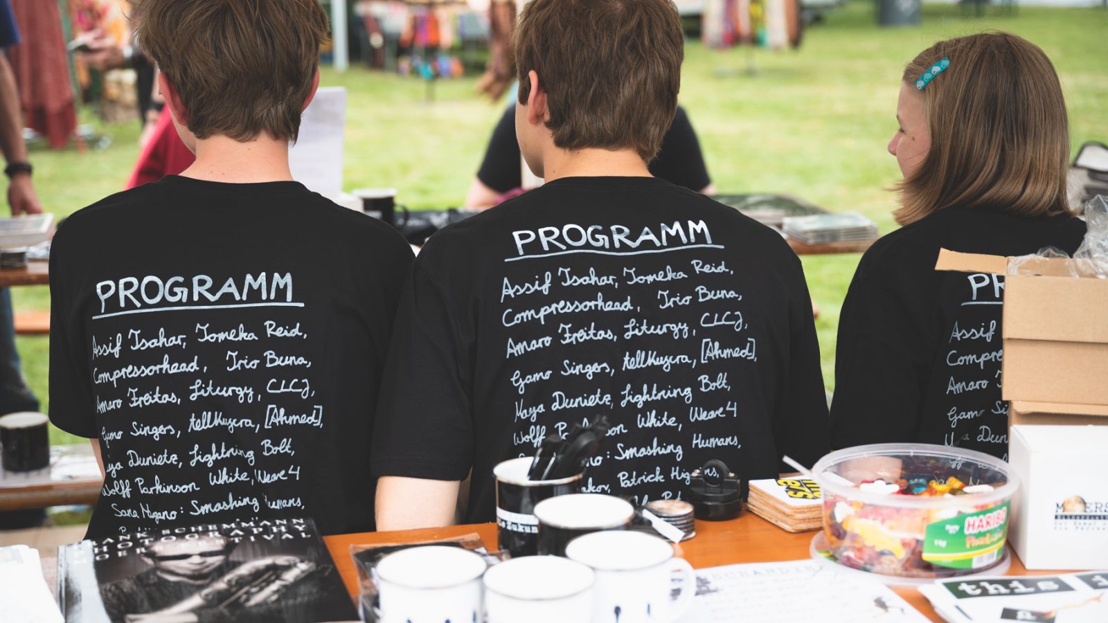 schwarz/weiß-Motiv: drei junge Volunteers sitzen auf einer Parkbank mit dem Rücken zum Betrachter, sie tragen das Festival T-Shirt 2022