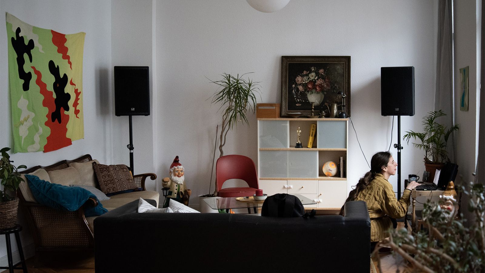 Wohnzimmer. Rechts hockt eine weiße Frau mit braunem Zopf und bedient einen Laptop. Im Hintergrund sieht man eine Kommode, ein Gemälde, eine Pflanze und Musikboxen. Links an der Wand hängt eine buntes Tuch über einem Sofa.