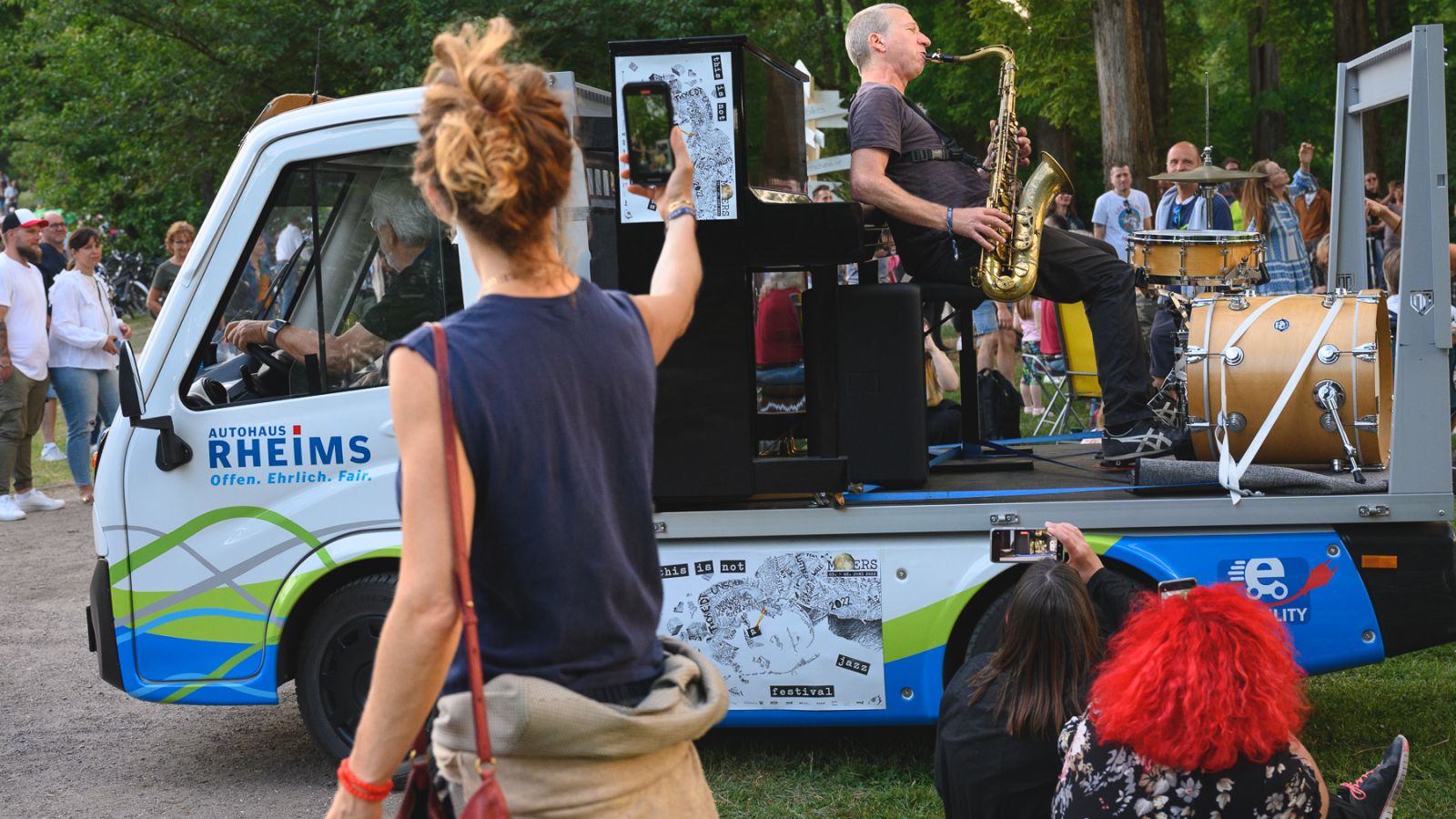 Ein Saxophonist sitzt auf einer Ladepritsche von einem kleinen Autotransporter und spielt Musik. Überall stehen Menschen, gucken und hören.