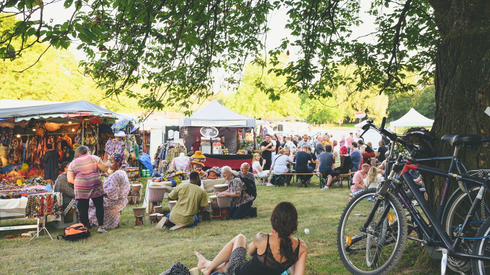 Blick ins rege Treiben vom Festivaldorf. Im Vordergrund liegt eine junge Frau auf der Decke und blickt auf die Verkaufsbuden. Am Rand sind Fahrräder an einem Baum abgestellt.