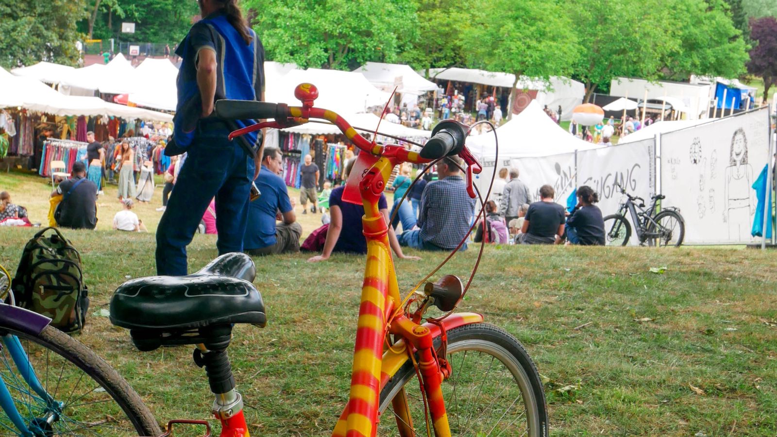 Ein buntgemaltes Fahrrad, abgestellt. Im Hintergrund ist reges Treiben auf dem Festivaldorf zu sehen.