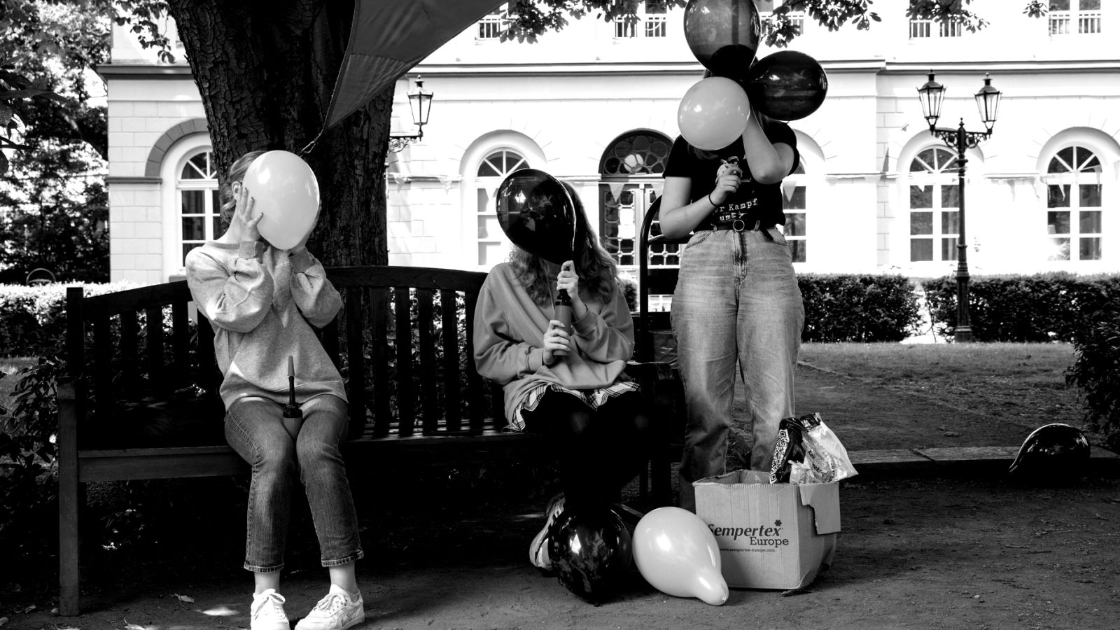 schwarz/weiß-Motiv: junge Volunteers sitzen auf einer Parkbank unter einem Baum und blasen Luftballons auf. Im Hintergrund ist das Marinsstift in Moers zu sehen.