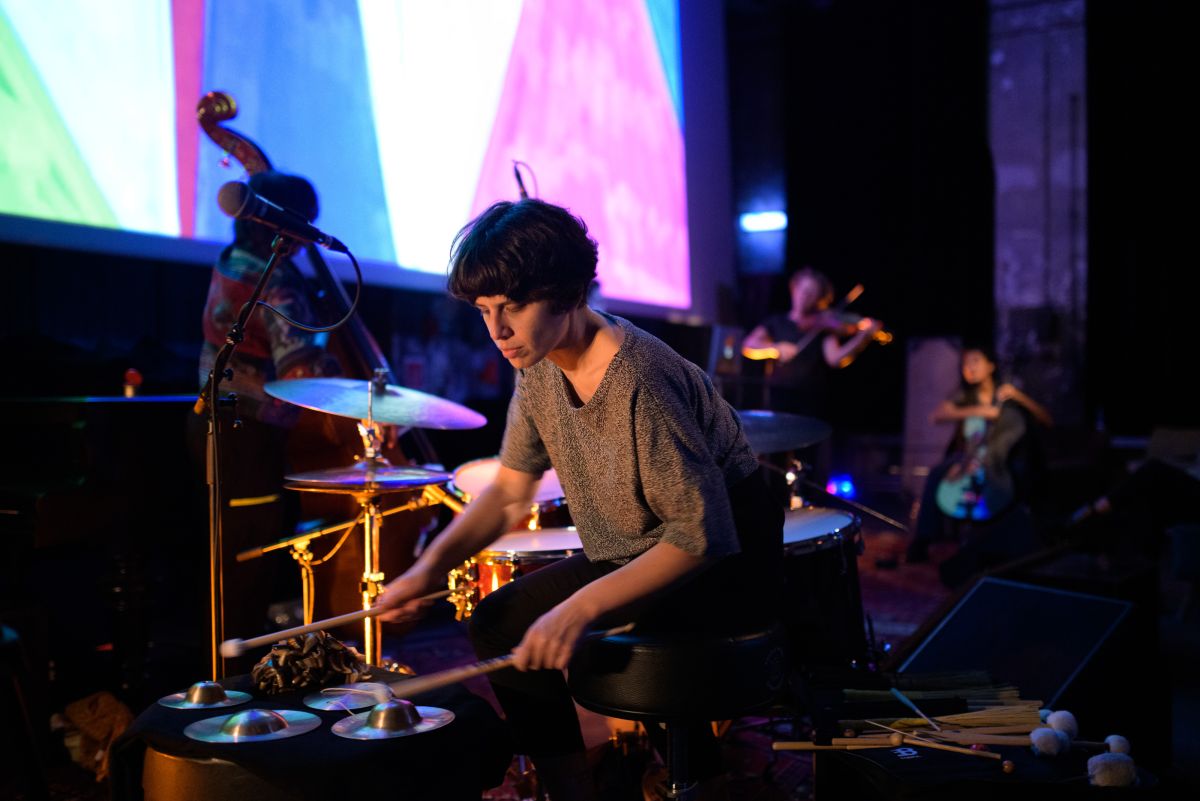 Eine Frau mit schwarzen Haaren, die auf Perkusssionsinstrumenten spielt. Im Hintergrund unterscharf weitere Musiker*innen