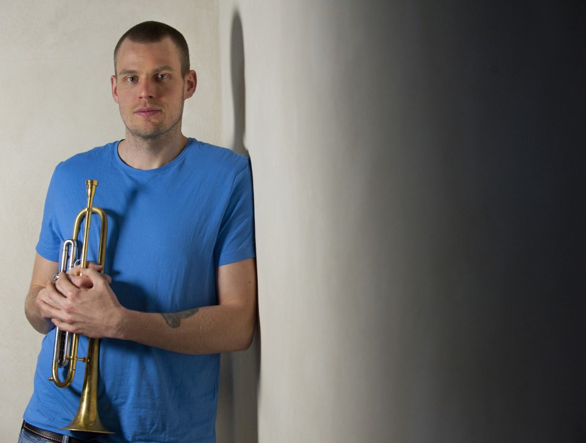 Mann mit kurzrasierten Haaren und blauem T-Shirt lehnt an einer Wand. In der Hand hält er eine Trompete